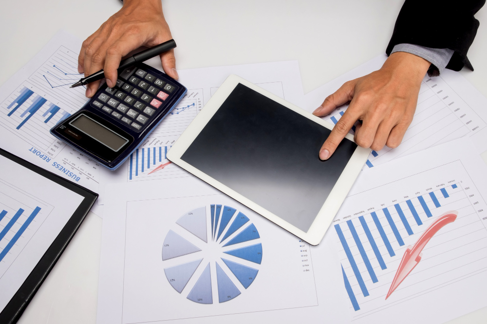 Mãos de profissional utilizando calculadora e tablet sobre mesa com relatórios financeiros, gráficos de barras e gráfico de pizza durante análise de indicadores de desempenho empresarial.