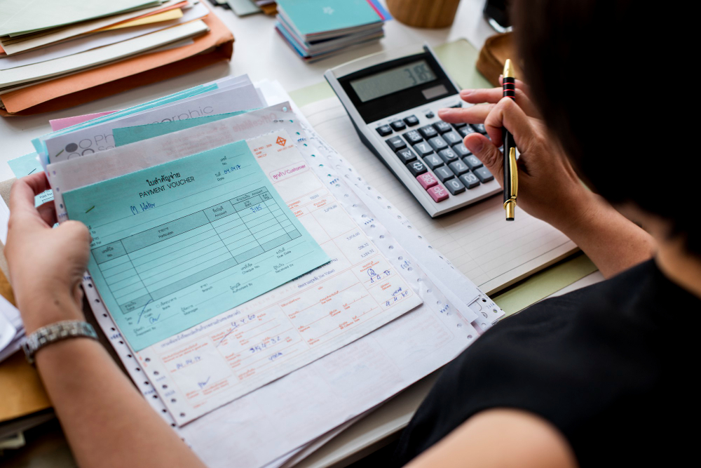 Pessoa analisando documentos fiscais e utilizando calculadora para organizar informações tributárias em mesa de trabalho.