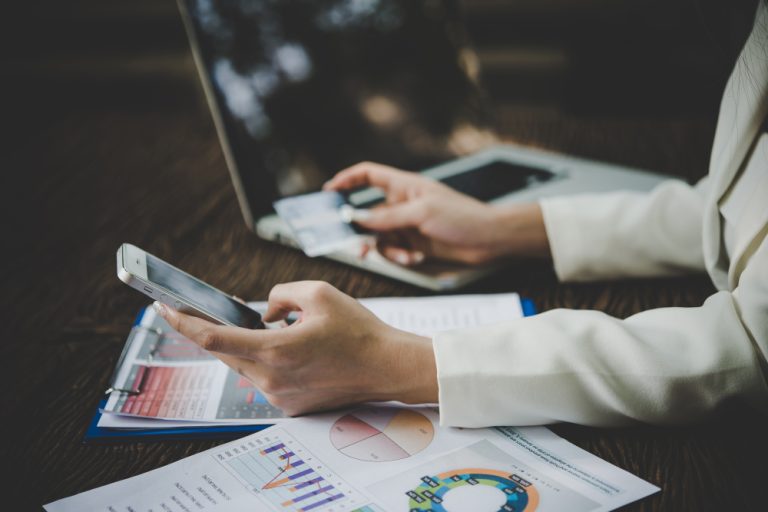 Pessoa utilizando smartphone e cartão bancário enquanto analisa gráficos financeiros e documentos ao lado de um notebook.