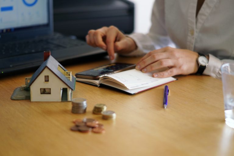 Pessoa utilizando calculadora para calcular tributos, com moedas e miniatura de casa sobre a mesa ao lado de notebook.