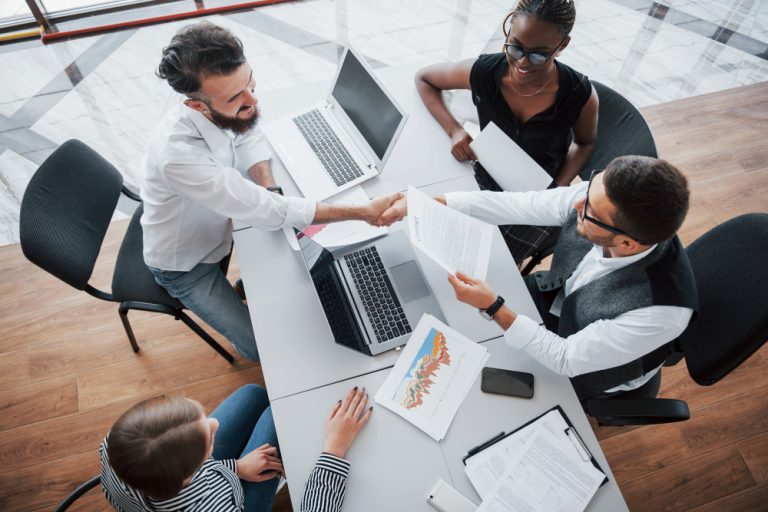 Profissionais reunidos em torno de uma mesa analisando documentos e gráficos, com aperto de mãos durante uma negociação empresarial.